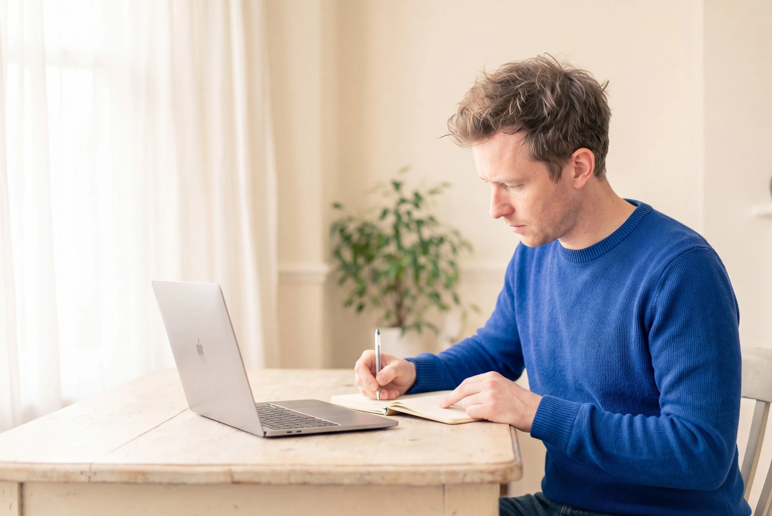 A man at a kitchen table, writing in a notebook beside his laptop, while the receptionist takes the call